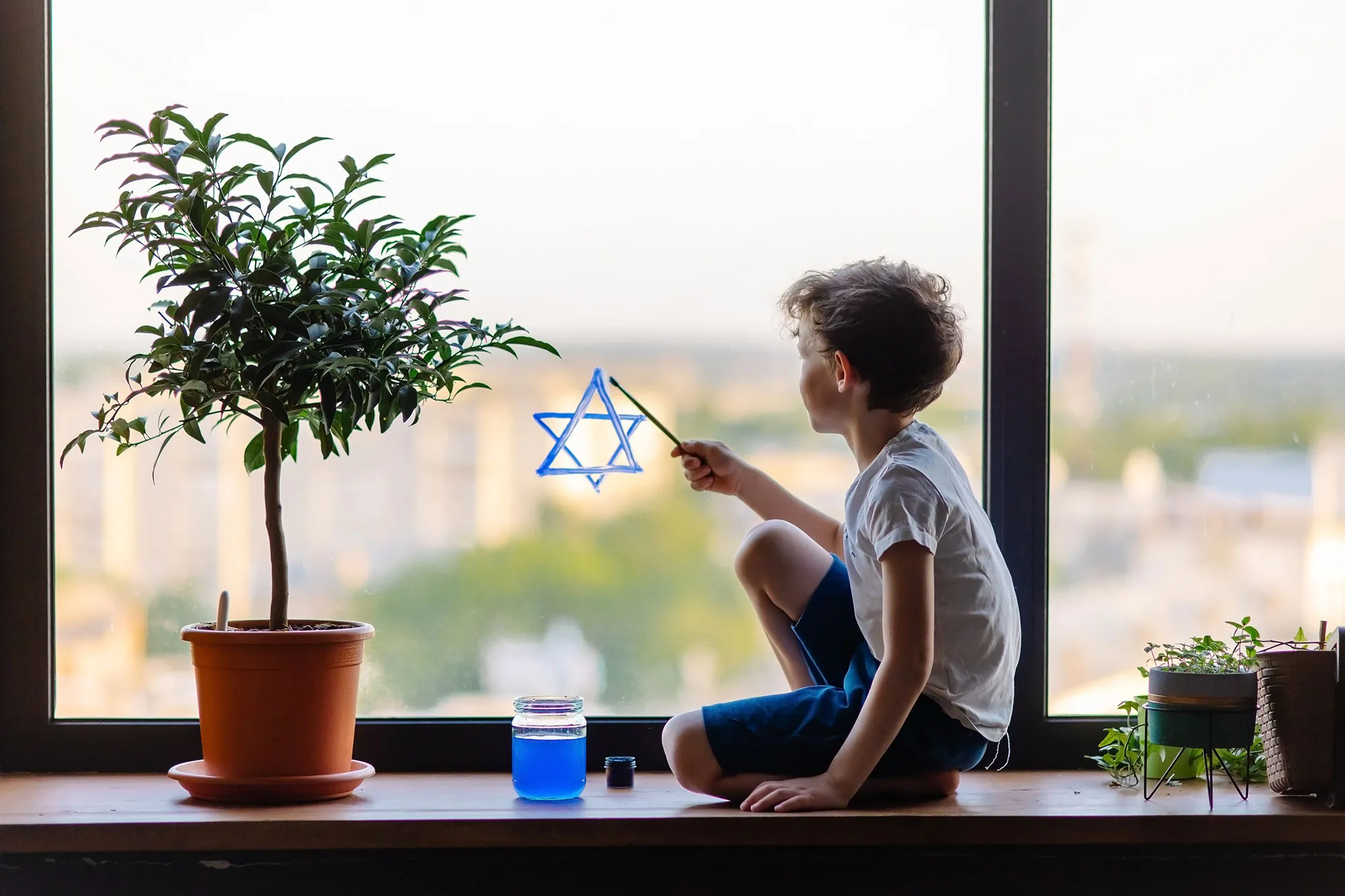 Junge sitzt auf Fensterbank und malt blauen Davidstern an Fenster