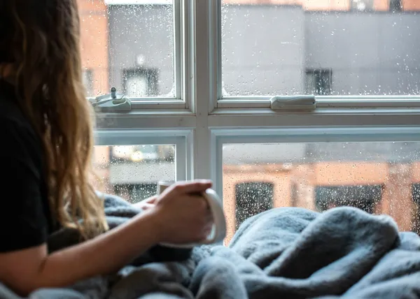 Quelle: Amin Hasani/Unsplash Frau sitzt mit Decke und Tasse am Fenster und schaut hinaus