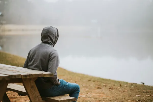Quelle: Amy Tran/Unsplash Mann im grauen Kapuzenpullover sitzt auf einer Bank und blickt auf einen Fluss im Nebel