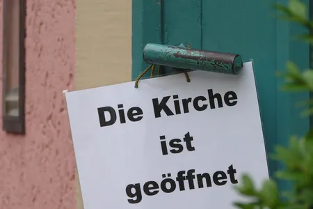 Kirchentür geöffnet, an der Türklinke hängt ein Schild mit Aufschrift "Die Kirche ist geöffnet"