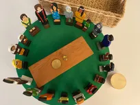 Godly Play Material zu dem guten Hirten und die weltweite Einheit der Christen: Auf einem grünen Holzkreis stehen 20 Holzfiguren, 1 Holztisch und ein Kelch + Teller aus Metall (in einer Schachtel)
