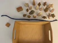 Godly Play Material zu Jakob: In einer kleinen offenen Holzkiste befinden sich 1 blauer Wollfaden, 2 Holzklötze, 1 Stein, 6 Holzfiguren (Erwachsene) und 12 Holzfiguren (Kinder)