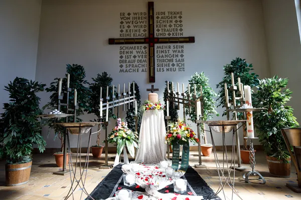Blick in Kapelle auf das Kreuz an der Wand,in der Mitte darunte eine weißverhülltr Stele mit Urne und Blumenschmuck, rechts und links mehrere Kerzenständer mit weißen Kerzen und die Osterkerze.